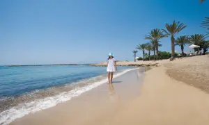 Dziewczynka w białej sukience i niebieskim kapeluszu spaceruje po piaszczystej plaży w Pafos. Delikatne fale obmywają jej stopy.