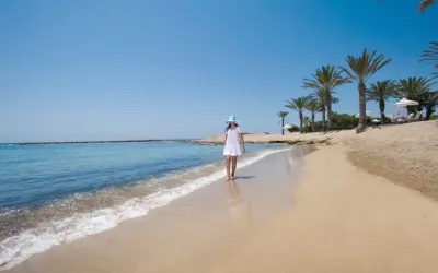 Dziewczynka w białej sukience i niebieskim kapeluszu spaceruje po piaszczystej plaży w Pafos. Delikatne fale obmywają jej stopy.