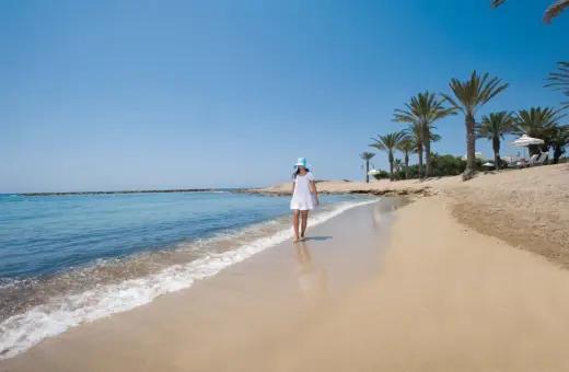 Dziewczynka w białej sukience i niebieskim kapeluszu spaceruje po piaszczystej plaży w Pafos. Delikatne fale obmywają jej stopy.