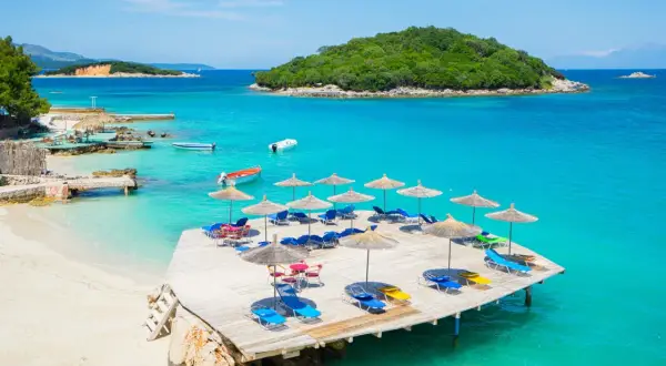 Słoneczny dzień na plaży Ksamil. Drewniany pomost z leżakami i parasolami, turkusowa woda i zielona wyspa w tle.