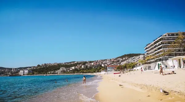 Słoneczna plaża w Grecji, turkusowe morze, białe budynki na wzgórzu. Idealne miejsce na wakacje, gdzie grecja ceny są przystępne.