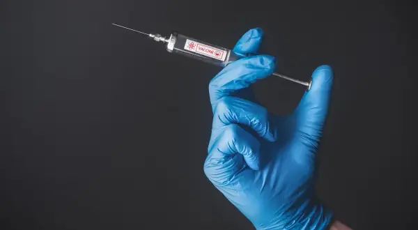 Ręka w niebieskiej rękawiczce trzyma strzykawkę z napisem VACCINE. To symbol szczepień, być może w kontekście Tunezji.
