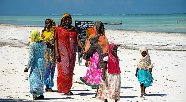 Kobiety i dzieci w kolorowych strojach spacerują po plaży Zanzibaru. Przed wyjazdem warto pomyśleć o zanzibar szczepienia.