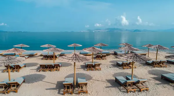 Puste leżaki i parasole na piaszczystej plaży, idealne na spokojny wypoczynek. W tle błękitne morze i góry.