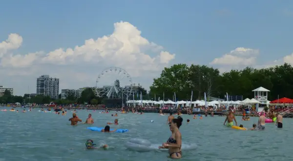 Ludzie kąpią się i relaksują na **Balaton plaża**. W tle widać diabelski młyn i budynki.