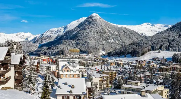 Zimowy krajobraz z ośnieżonymi górami i lasami, gdzie jest Davos. W dole widać zabudowania i hotel.