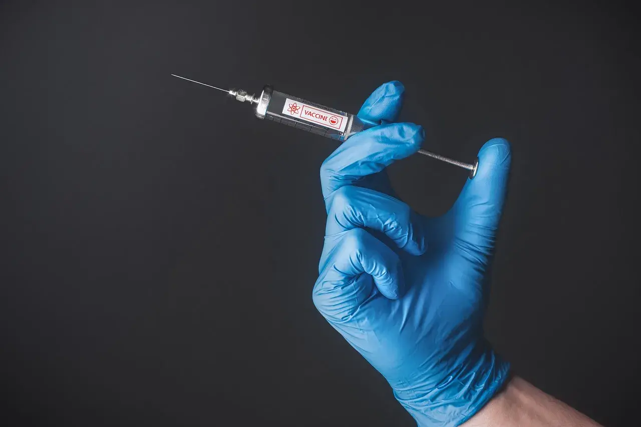 Ręka w niebieskiej rękawiczce trzyma strzykawkę z napisem VACCINE. To symbol szczepień, być może w kontekście Tunezji.