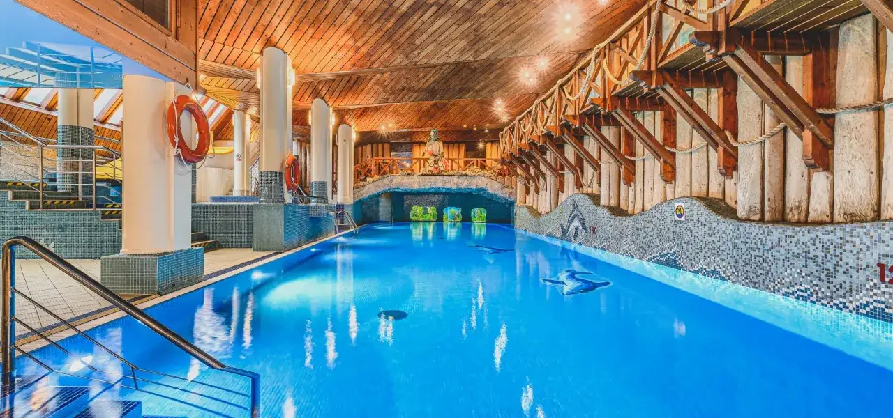 Basen w hotelu Belvedere Resort & Spa z drewnianym sufitem i kamiennymi zdobieniami. Woda jest błękitna, a na dnie widać mozaiki z delfinami.